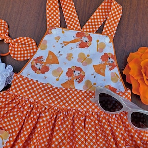 May include: Orange and white gingham overall dress with a white bodice featuring a repeating pattern of cartoon girls with orange hair and butterfly designs.