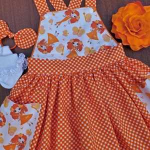 May include: Orange and white gingham dress with a white bodice featuring a repeating pattern of orange haired girls with butterfly wings. The dress has a bib style top with adjustable straps.