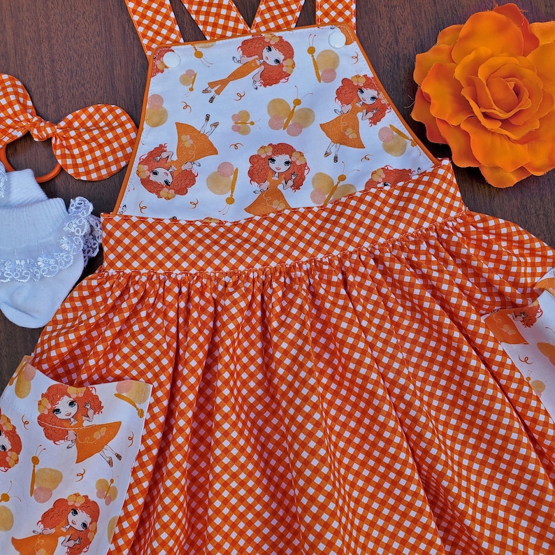 May include: Orange and white gingham dress with a white bodice featuring a repeating pattern of orange haired girls with butterfly wings. The dress has a bib style top with adjustable straps.