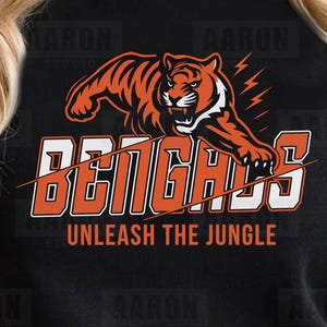 May include: Black sweatshirt featuring an orange and white tiger graphic with the word "BENGALS" and the phrase "UNLEASH THE JUNGLE". The design includes lightning bolts and is likely a sports-themed apparel item.