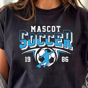 May include: Black t-shirt featuring a blue and white "MASCOT SOCCER" design. The graphic includes a soccer ball and player silhouette, with the numbers "19" and "86". The shirt has a crew neck and rolled-up sleeves.