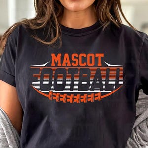 Pode incluir: Camiseta preta com as palavras "MASCOT FOOTBALL" em laranja e branco. O design apresenta uma forma de bola de futebol com o texto dentro. A camisa é de estilo casual.