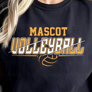 Pode incluir: Camiseta preta com as palavras "MASCOT VOLLEYBALL" em letras douradas e brancas. Um gráfico de voleibol está abaixo do texto. A camiseta é usada por uma pessoa.