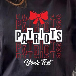 Può includere: T-shirt nera con la scritta "PATRIOTS" in un design bianco e rosso, sormontata da un fiocco rosso. La maglietta include anche il testo "Your Text" in basso. Il design è probabilmente per una squadra sportiva o un tema patriottico.