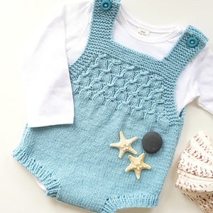 以下が含まれることがあります： 白い肌着を着た薄い青色のニットのベビーロンパース。ロンパースはテクスチャのあるパターンとストラップに2つの青いボタンが付いています。ロンパースの前には、ヒトデが2つと灰色の石が1つあります。