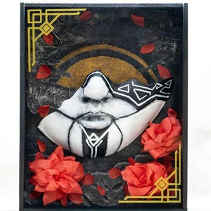 Op de afbeelding: Een ingelijst kunstwerk met een wit masker met zwarte geometrische ontwerpen, tegen een donkere achtergrond met gouden accenten en rode papieren bloemaccenten. Het kunstwerk is ingelijst in een zwarte doos.