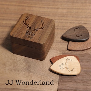 Könnte beinhalten: Eine Holzbox und Gitarrenplektren mit eingravierten Designs. Die Box ist quadratisch mit einem Deckel und zeigt ein Hirschgeweih-Design sowie den Namen "Eric" mit dem Jahr "2024". Mehrere Gitarrenplektren sind sichtbar, einige mit Initialen und andere mit ähnlichen Gravuren.