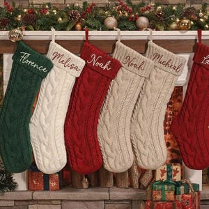 Op de afbeelding: Een gezellige kerstscène met zes kabelgebreide kousen in groen, wit, rood en crème, elk gepersonaliseerd met een naam in elegant schrift. De kousen hangen boven een open haard versierd met feestelijk groen en twinkelende lichtjes.