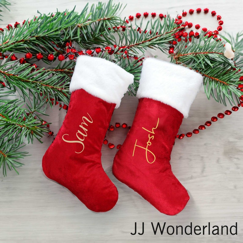 May include: Two red Christmas stockings with white fur trim and gold embroidered names, "Sam" and "Josh", on a woodgrain background with red and white beaded garland.