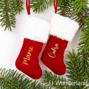 May include: Two red Christmas stockings with white faux fur trim, hanging from a green pine branch. The stockings are personalized with the names "Marie" and "Calbra" in gold lettering.