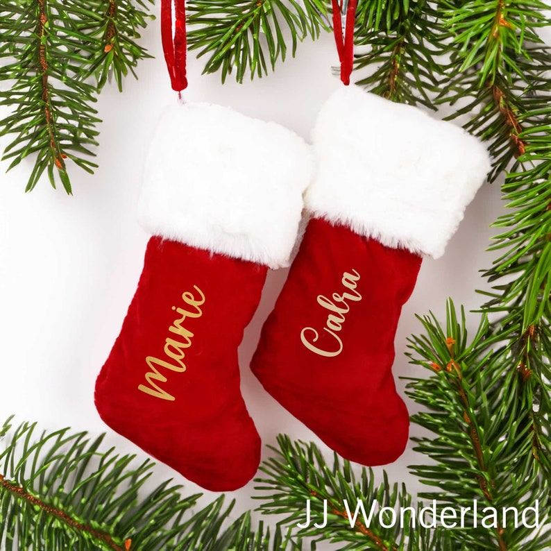 May include: Two red Christmas stockings with white faux fur trim, hanging from a green pine branch. The stockings are personalized with the names "Marie" and "Calbra" in gold lettering.
