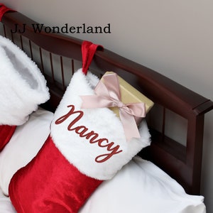 Puede incluir: Calcet&iacute;n de Navidad rojo y blanco con un pu&ntilde;o de piel sint&eacute;tica blanca, personalizado con el nombre "Nancy" en bordado rojo. Una caja de regalo dorada con una cinta rosa est&aacute; dentro del calcet&iacute;n.