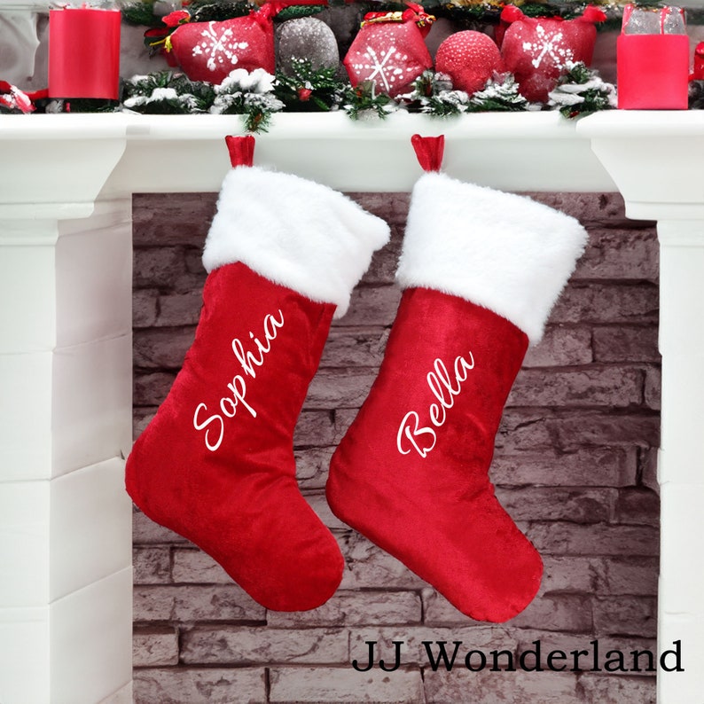 May include: Two red velvet Christmas stockings with white faux fur trim, hanging from a white fireplace mantel. The stockings are personalized with the names "Sophia" and "Bella" in white cursive lettering.