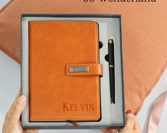 Könnte beinhalten: Ein braunes, ledergebundenes Tagebuch mit einem Stift in einer Geschenkbox. Das Tagebuch trägt den Namen "KELVIN" auf der Vorderseite und ein silbernes Namensschild mit demselben Namen. Die Box ist schwarz mit einem grauen Innenraum.