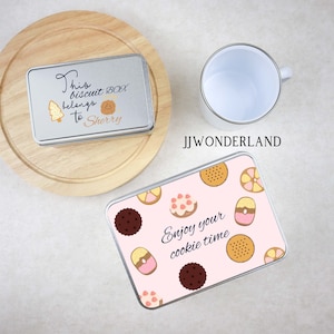 May include: Two silver metal cookie tins. One tin has a pink background with a pattern of colorful cookies and the text "Enjoy your cookie time". The other tin has a silver background with the text "This biscuit box belongs to Sherry".