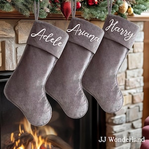 Puede incluir: Tres calcetines de Navidad de terciopelo gris con nombres bordados en blanco: Adele, Ariana y Harry. Los calcetines están colgados en la repisa de la chimenea.