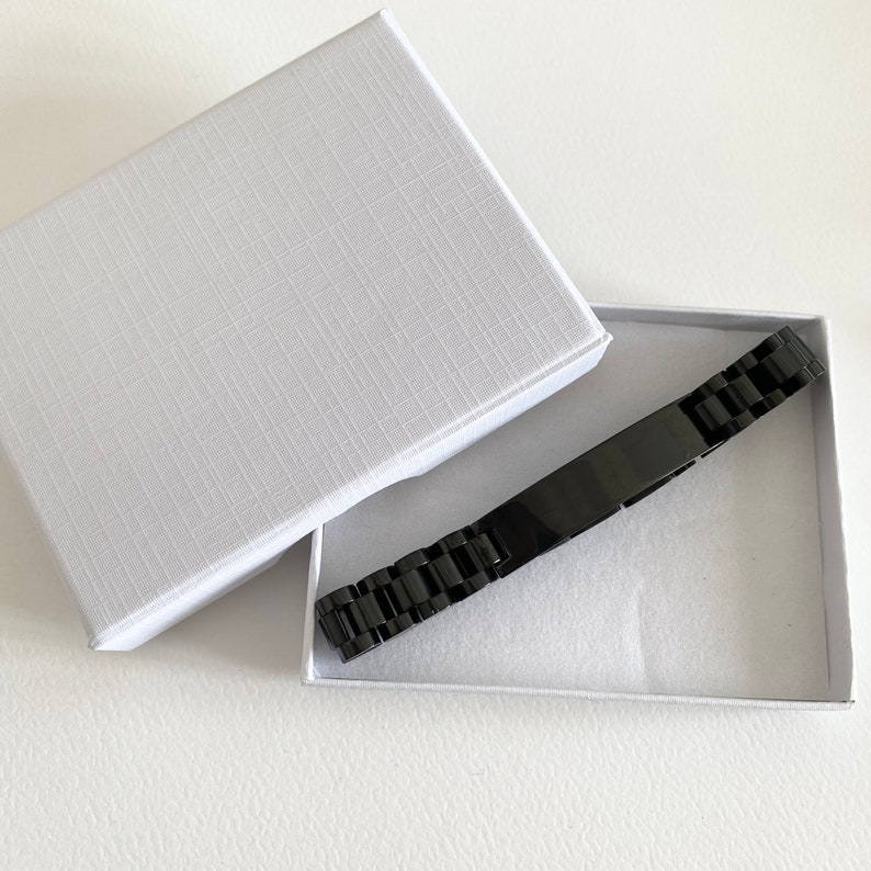 May include: A black metal bracelet with a rectangular plate, in a white box with a textured lid.