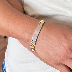 May include: A silver and gold bracelet with a rectangular nameplate. The bracelet has a watch-style band.
