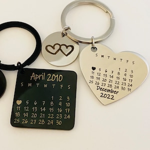 May include: Two silver keychains with engraved calendars. One keychain features a square calendar with the month of April 2010, and the other keychain features a heart-shaped calendar with the month of December 2022. Both keychains have two intertwined hearts engraved on a circular tag.