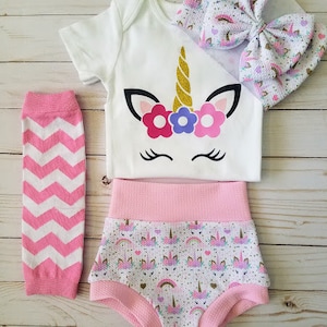 May include: A white onesie with a unicorn design, pink and white leggings, and a pink and white diaper cover with a unicorn print. The onesie has a gold horn and pink and purple flowers on the unicorn's head.