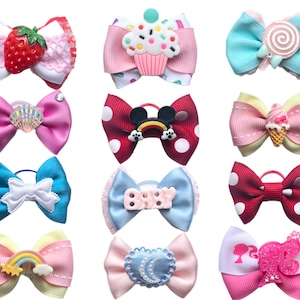 May include: Twelve colourful hair bows with various designs, including a cupcake, a lollipop, a rainbow, a seashell, a Mickey Mouse head, and a Barbie doll. The bows are made of ribbon and have a variety of embellishments.