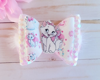 May include: A white and pink hair bow with a white cat cartoon character in the center. The bow is made of fabric with a white background and pink polka dots. The bow is trimmed with white fur.