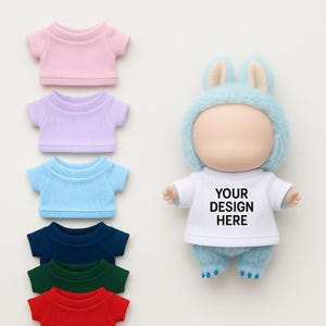 Könnte beinhalten: Miniatur-T-Shirts in Rosa, Lila, Hellblau, Marineblau, Grün und Rot. Eine Puppe trägt ein weißes T-Shirt mit dem Text "YOUR DESIGN HERE" und einen blauen Hasen-Hut und -Hose.