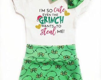 May include: A white baby onesie with the text "I'm so cute even the Grinch wants to steal me!" in pink and green lettering with gold glitter hearts. The onesie is paired with green shorts featuring a repeating pattern of the Grinch's face. A green Grinch-themed bow is on top of the onesie.
