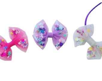 May include: Three hair bows with purple, pink, and white tulle fabric. Each bow is decorated with colorful sequins and glitter.