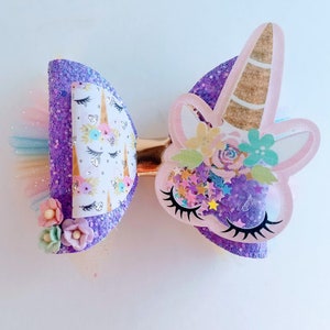 May include: Purple and pink glitter unicorn hair bow with a white unicorn face, flowers, and glitter stars. The bow has a gold clip.