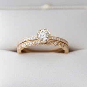 May include: A gold ring set consisting of a solitaire ring with a round center stone and a matching scalloped band. Both rings feature delicate milgrain detailing and small embedded stones along the bands, presented in a white box.