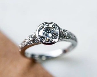 May include: A silver ring with a large, round, clear gemstone at the center. The band is detailed with small, decorative embellishments. The ring is set against a neutral background, highlighting its intricate design and sparkle.
