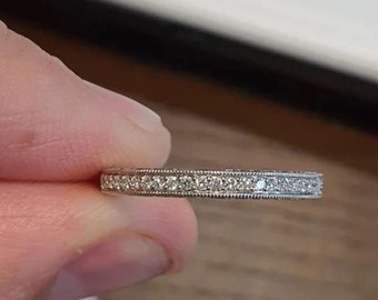 May include: A delicate, silver-colored ring with a continuous row of small, clear gemstones. The ring is held between fingers, showcasing its intricate design and fine details. The ring's band features a textured border.