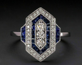 May include: A silver ring with a geometric design. The ring features a central row of three round diamonds, surrounded by blue sapphires and smaller diamonds. The ring has a detailed, Art Deco style.
