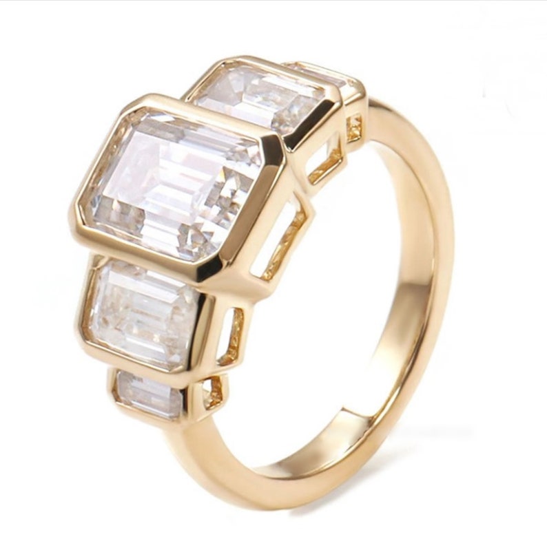 May include: A gold ring with three rectangular, clear gemstones. The gemstones are set in a gold band with a simple, elegant design. The ring is displayed against a white background.