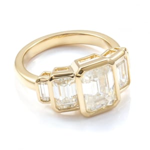 May include: A gold ring with three emerald-cut diamonds. The ring has a polished gold band. The diamonds are set to showcase their clarity and brilliance. The diamonds are arranged in a row, creating a classic and elegant design. The ring is a piece of jewellery.