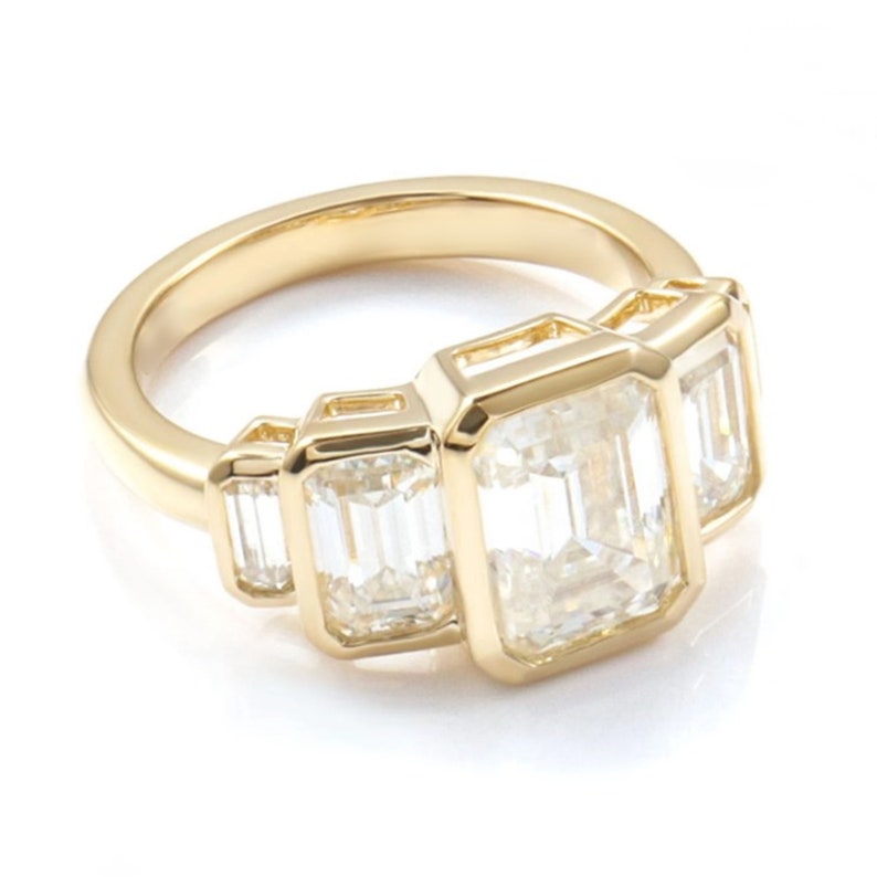 May include: A gold ring with three emerald-cut diamonds. The ring has a polished gold band. The diamonds are set to showcase their clarity and brilliance. The diamonds are arranged in a row, creating a classic and elegant design. The ring is a piece of jewellery.