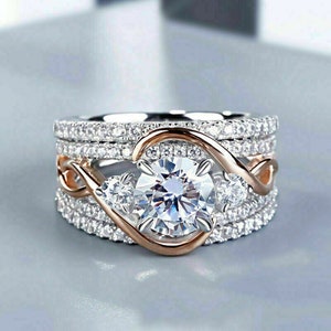 May include: A three-piece ring set featuring a large, round, clear center stone, flanked by two smaller stones. The rings have a silver band with small, clear stones and a rose gold swirl design.