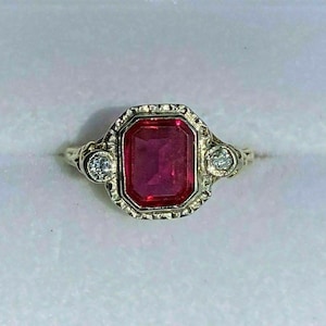 Puede incluir: Un anillo de estilo vintage con una gran gema rectangular de color rojo rubí como pieza central. El rubí está rodeado por un engaste decorativo en tono dorado, con una pequeña gema redonda y transparente a cada lado. El anillo está sobre un fondo blanco.