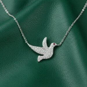 Op de afbeelding: Een zilveren ketting met een duifhanger, bezet met kleine, sprankelende stenen. De duif heeft een rode edelsteen oog en hangt aan een fijne ketting. De ketting wordt getoond tegen een groene achtergrond.