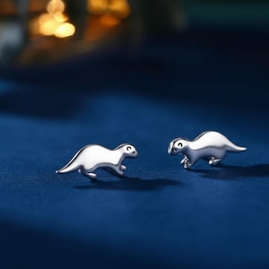 Pode incluir: Um par de brincos prateados em forma de pequenos animais estilizados. Cada brinco apresenta um corpo curvo, pernas pequenas e um pequeno olho escuro. Num fundo azul escuro.