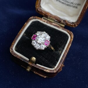 May include: A vintage ring with a central round diamond surrounded by smaller diamonds and two square rubies. The ring is set in a gold band and displayed in a brown leather jewelry box with a dark blue velvet interior.