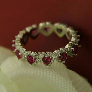 May include: A silver ring with a series of heart-shaped, ruby-colored gemstones. Each heart is surrounded by smaller, clear gemstones. The ring is set against a red background and a white fabric.