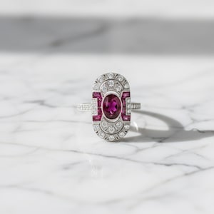 May include: An Art Deco-style ring with a large oval ruby center stone, surrounded by smaller rubies and diamonds. The ring has a silver band and a rectangular face with intricate detailing. The gemstones are set in a geometric pattern.