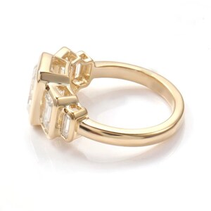 May include: A gold ring featuring a large, rectangular central gemstone flanked by smaller rectangular stones. The ring has a polished finish and is set against a white backdrop. The design is modern and elegant, suitable for special occasions.