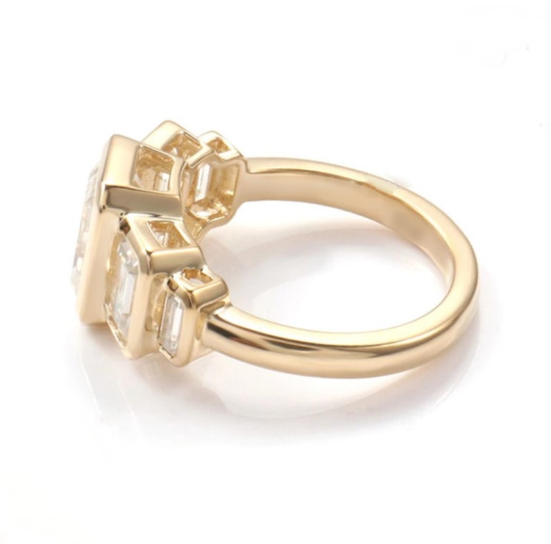 May include: A gold ring featuring a large, rectangular central gemstone flanked by smaller rectangular stones. The ring has a polished finish and is set against a white backdrop. The design is modern and elegant, suitable for special occasions.