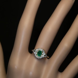 May include: A gold ring featuring an oval emerald center stone surrounded by a halo of small, round diamonds. The ring is set on a gold band and is displayed on a hand against a black background.
