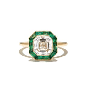 Może przedstawiać: Złoty pierścionek z dużą, kwadratową, przezroczystą kamieniem szlachetnym, otoczonym szmaragdowo-zielonymi kamieniami o szlifie bagietowym. Pierścionek ma delikatną obrączkę i otoczkę zielonych kamieni wokół centralnego klejnotu. Na białym tle.