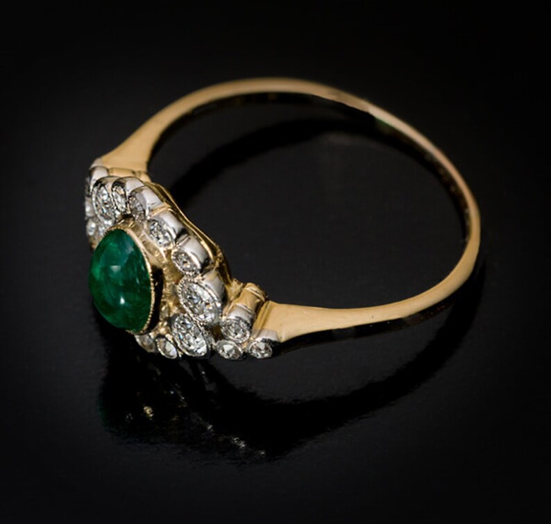May include: A gold and silver ring with a teardrop-shaped emerald center stone. The emerald is surrounded by small, round diamonds. The ring has a delicate, curved band.