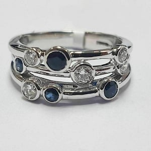 Puede incluir: Un anillo de plata con tres bandas, cada una adornada con piedras redondas alternas. Las piedras preciosas incluyen diamantes claros y zafiros azul oscuro, creando un diseño brillante. El anillo se muestra sobre un fondo blanco.